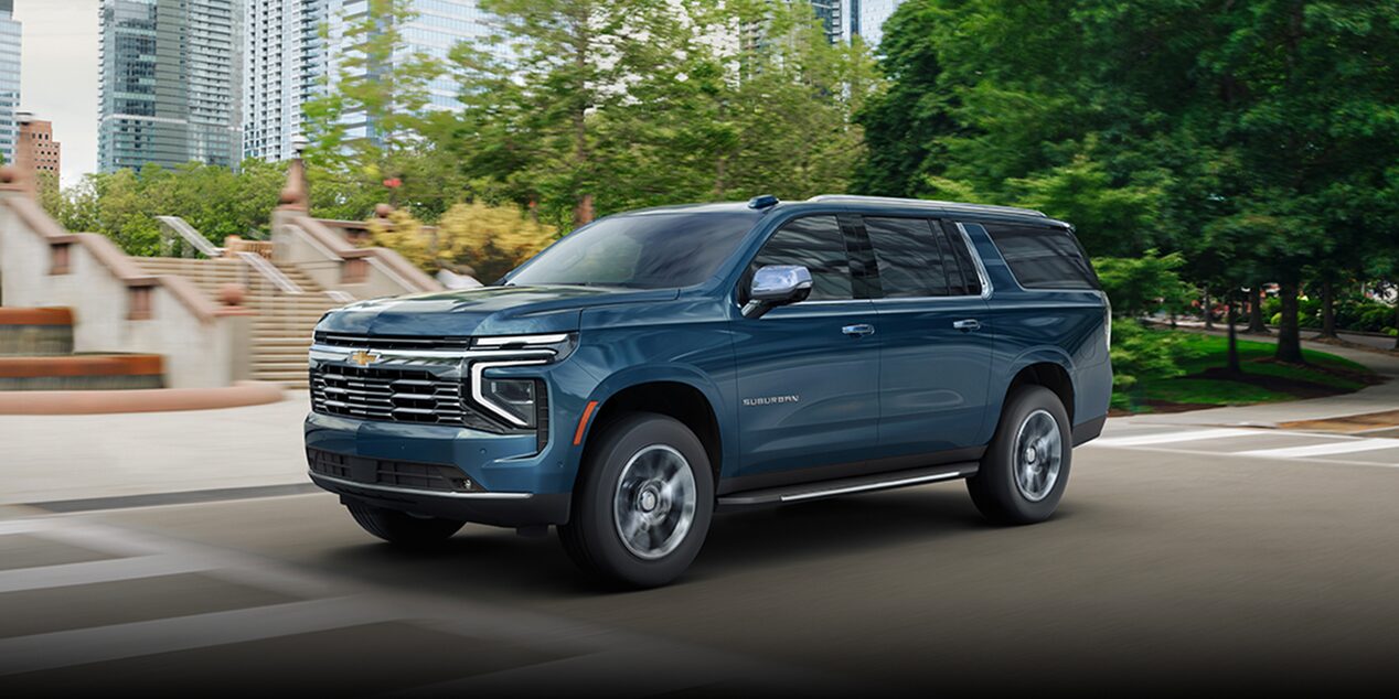 Driving image with front three quarters view of 2025 Chevrolet Suburban Premier in city surrounded by trees