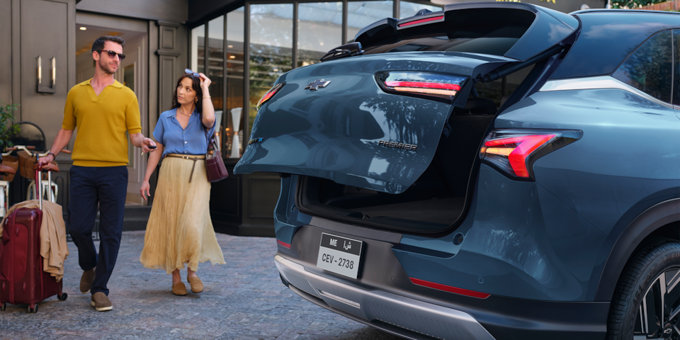 A couple walking with their luggage towards a Dawn Blue 2026 Chevrolet Captiva EV while opening trunk and parked in front of a hotel in a city