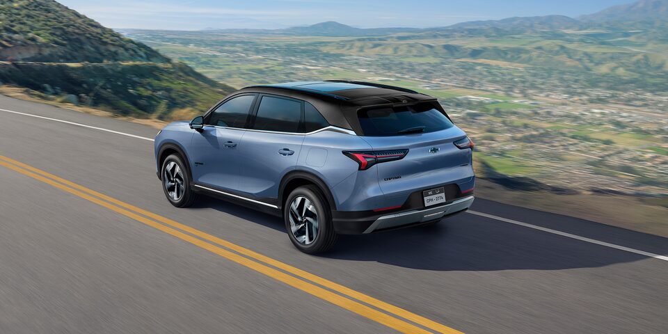 Rear Three Quarters of a Dawn Blue 2026 Chevrolet Captiva EV Driving Down a Mountain Road Overlooking a City Landscape