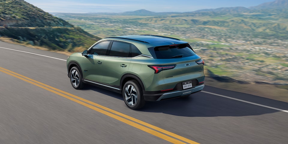 Rear Three Quarters of a Khaki Green 2026 Chevrolet Captiva PHEV Driving Down a Mountain Road Overlooking a City Landscape