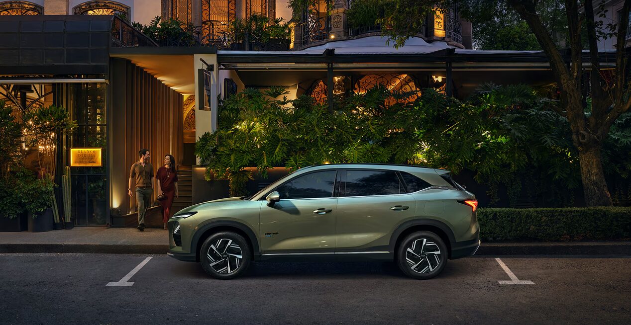 A couple walking towards Left of Khaki Green 2026 Chevrolet Captiva PHEV parked at night in front of a hotel