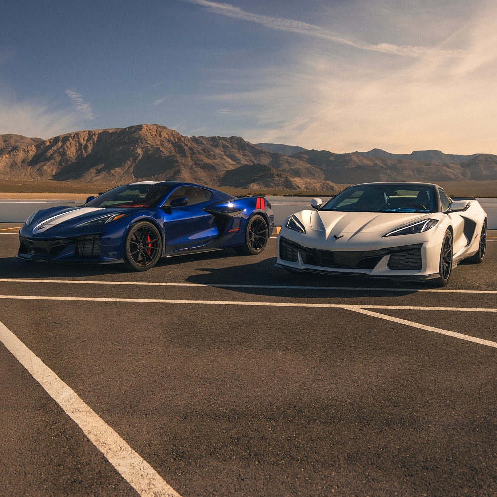 A Blue Chevrolet Corvette Grand Sport and a White Chevrolet Corvette Grand Sport Are Parked Side by Side on a Racetrack With Mountain Ridges in the Distance Under a Clear Sky.