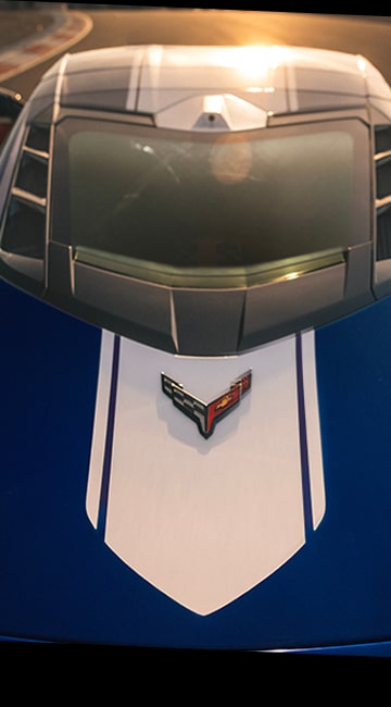 The Front Hood of a Blue Chevrolet Corvette Grand Sport With White Racing Stripes and the Corvette Emblem Is Shown Under Warm Sunset Light on a Racetrack.