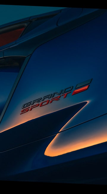 A Close‑Up View of the Grand Sport Badge on the Blue Chevrolet Corvette Grand Sport Shows the Metallic Lettering and Red Accent Stripe Reflecting Warm Sunset Light.