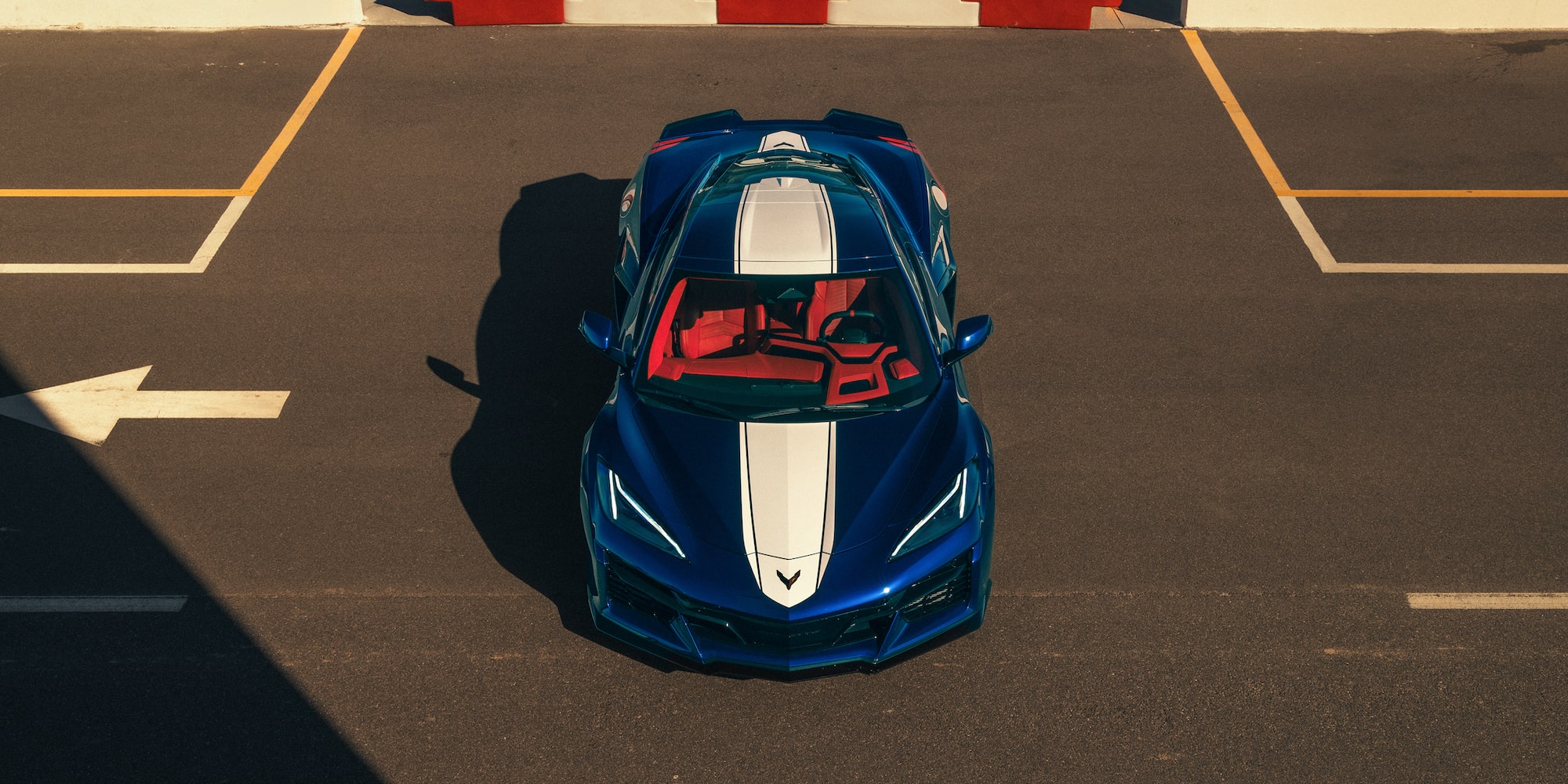 A Blue Chevrolet Corvette Grand Sport With White Racing Stripes Is Parked in a Sunlit Asphalt Lot With Shadows Cast Across the Ground.