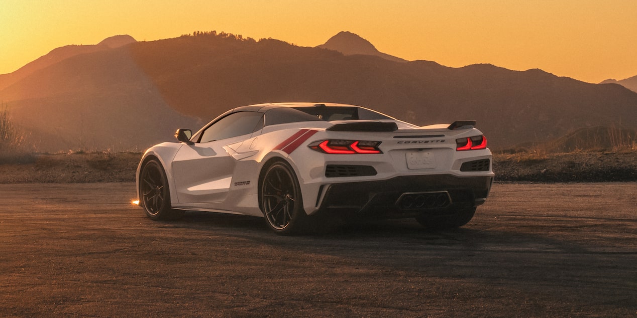 The Rear of a White Chevrolet Corvette Grand Sport With Quad Exhaust Outlets and Red Accent Stripes Is Shown Parked on Sunlit Dirt With Mountains Silhouetted at Sunset.