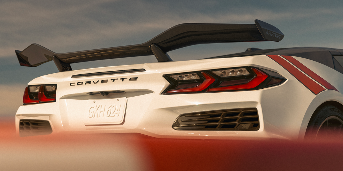 The Rear of a White Chevrolet Corvette Grand Sport With a Large Rear Wing and Red Accent Stripes Appears Above a Red‑And‑White Track Barrier in Warm Sunset Light.