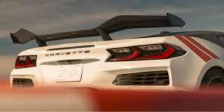 The Rear of a White Chevrolet Corvette Grand Sport With a Large Rear Wing and Red Accent Stripes Appears Above a Red‑And‑White Track Barrier in Warm Sunset Light.
