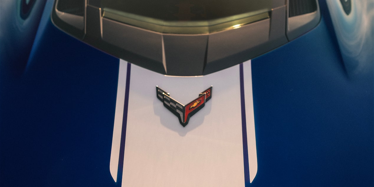 A Close View of the Front Hood of a Blue Chevrolet Corvette Grand Sport Shows White Racing Stripes and the Corvette Emblem Under Warm Sunlight.