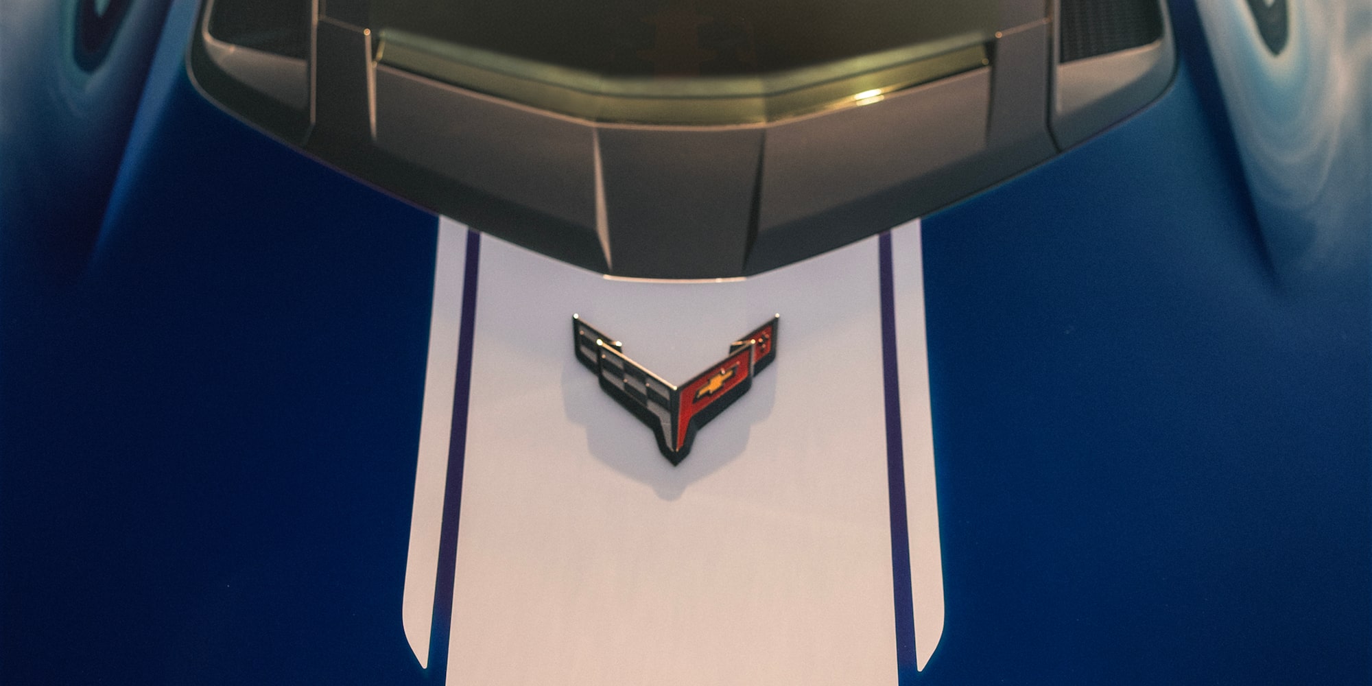A Close View of the Front Hood of a Blue Chevrolet Corvette Grand Sport Shows White Racing Stripes and the Corvette Emblem Under Warm Sunlight.