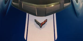 A Close View of the Front Hood of a Blue Chevrolet Corvette Grand Sport Shows White Racing Stripes and the Corvette Emblem Under Warm Sunlight.
