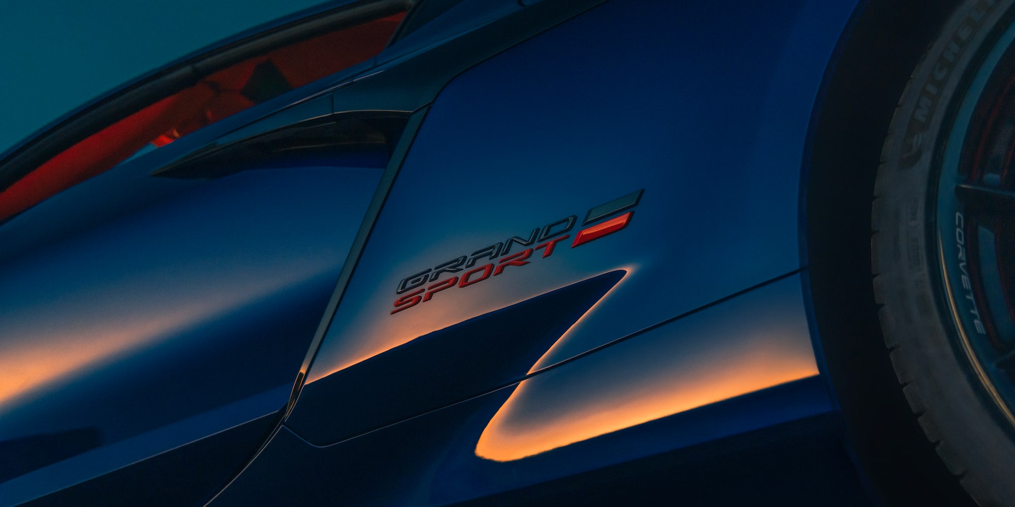 A Close View of the Front Hood of a Blue Chevrolet Corvette Grand Sport Shows White Racing Stripes and the Corvette Emblem Under Warm Sunlight.