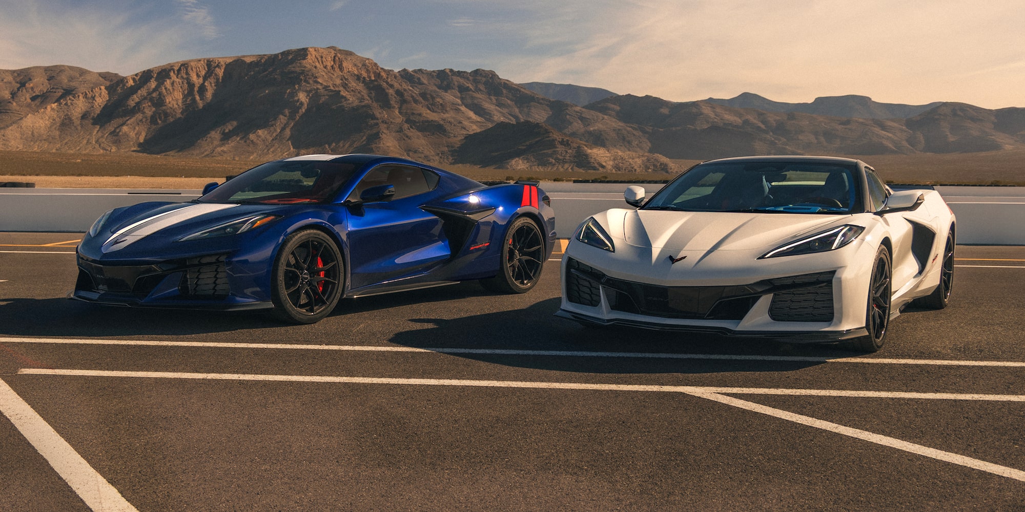 A Blue Chevrolet Corvette Grand Sport and a White Chevrolet Corvette Grand Sport Are Parked Side by Side on a Racetrack With Mountain Ridges in the Distance Under a Clear Sky.