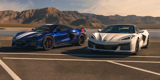 A Blue Chevrolet Corvette Grand Sport and a White Chevrolet Corvette Grand Sport Are Parked Side by Side on a Racetrack With Mountain Ridges in the Distance Under a Clear Sky.