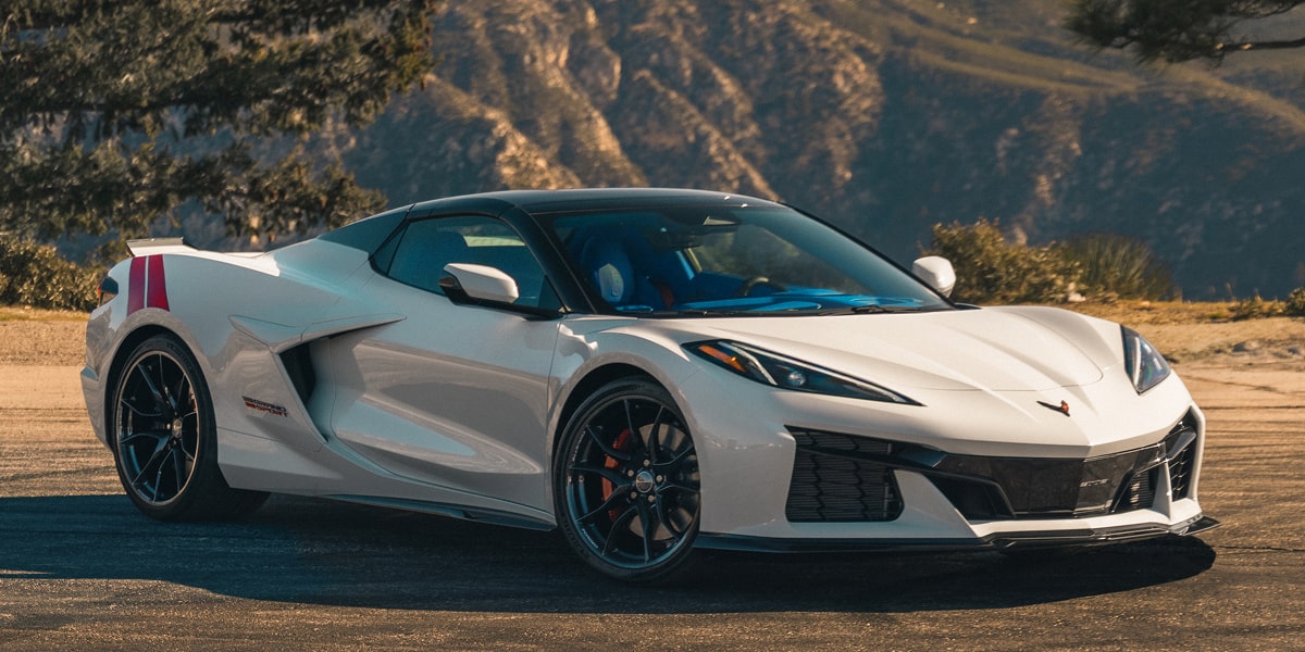 A White Chevrolet Corvette Grand Sport With Red Accent Stripes Is Parked Beside a Racetrack Barrier With Mountain Ridges in the Background.