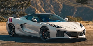 A White Chevrolet Corvette Grand Sport With Red Accent Stripes Is Parked Beside a Racetrack Barrier With Mountain Ridges in the Background.