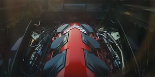 The Engine Bay of a Chevrolet Corvette Grand Sport Shows a Prominent Red Engine Cover Surrounded by Metallic and Black Components Under Soft Sunlight.