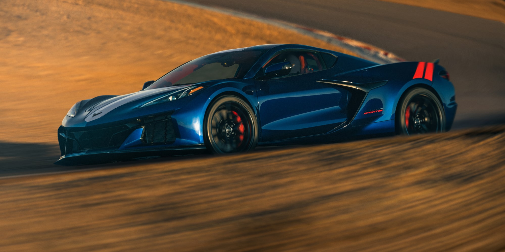 A Blue Chevrolet Corvette Grand Sport Speeds Through a Sand‑Covered Corner With Dust Billowing Around the Car in Warm Golden Light.