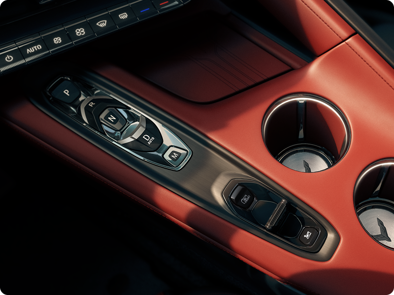 A Close‑Up View of the Center Console in a Chevrolet Corvette Grand Sport Shows Metallic Switch Controls Beside Two Circular Cupholders Set in Red Interior Trim.