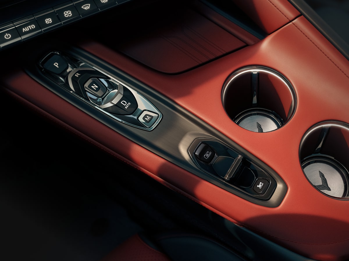 A Close‑Up View of the Center Console in a Chevrolet Corvette Grand Sport Shows Metallic Switch Controls Beside Two Circular Cupholders Set in Red Interior Trim.