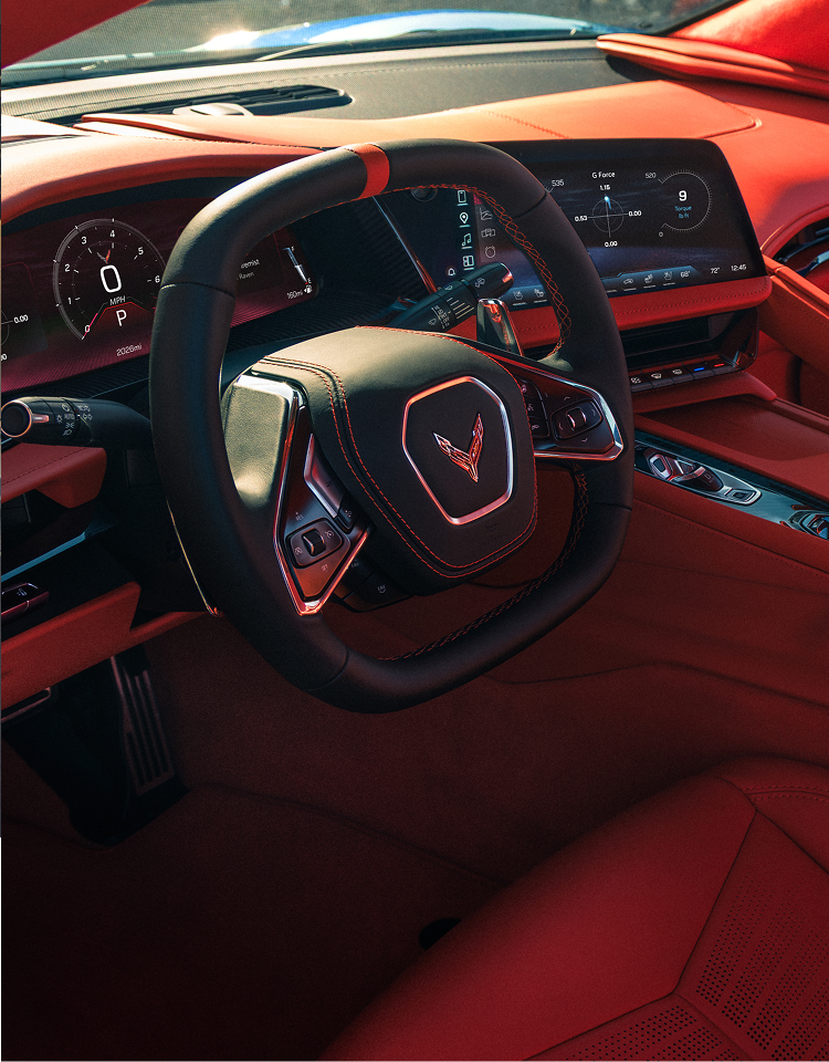 A Close‑Up of the Steering Wheel Inside a Chevrolet Corvette Grand Sport Shows the Corvette Emblem, Digital Instrument Cluster, and Red Interior Surfaces Illuminated by Sunlight.