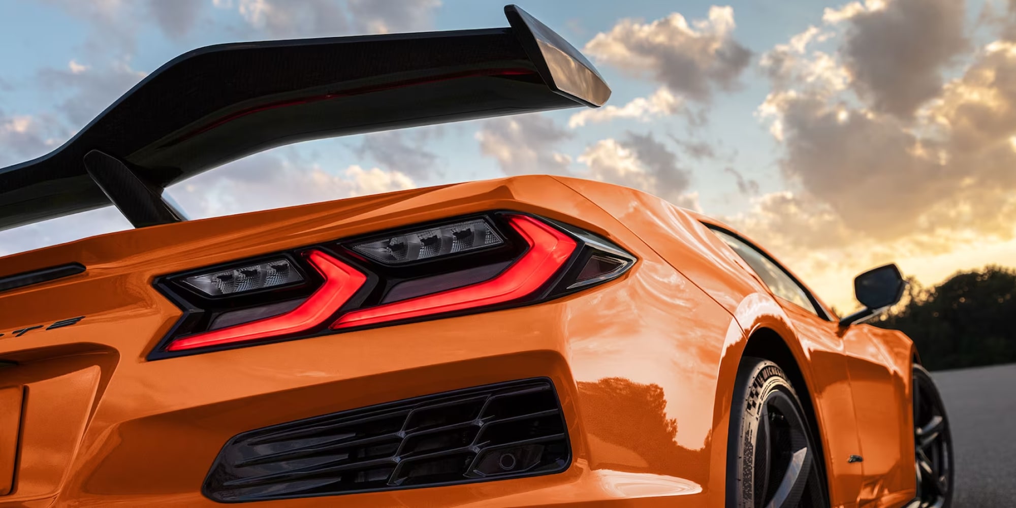 Close-up Rear Angle of an Orange 2027 Chevrolet Corvette Z06 with a Large Black Carbon Fiber Spoiler Against a Sunset Sky