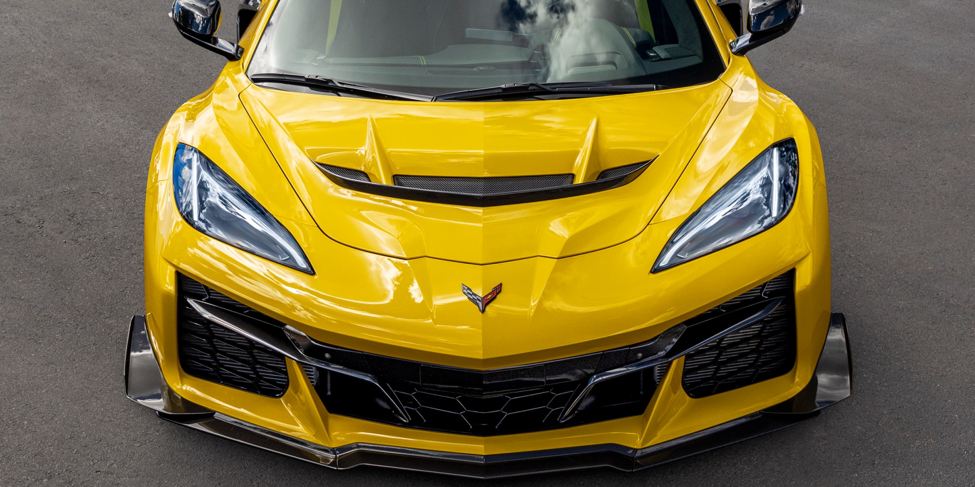 Un Chevrolet Corvette Grand Sport azul con franjas decorativas rojas circulando por una pista de carreras al lado de una barrera blanca, iluminado por la cálida luz del sol.