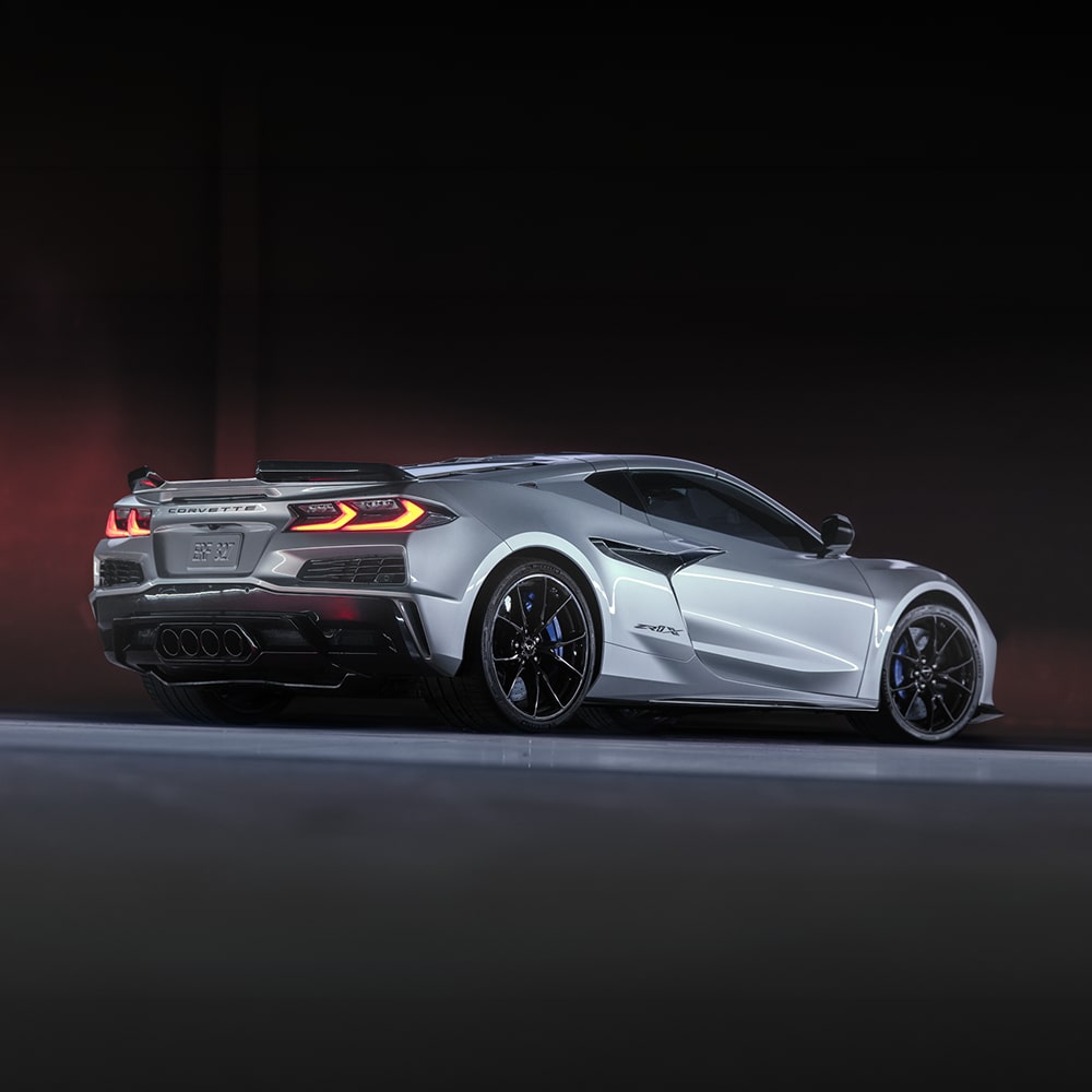 A Silver Chevy Corvette ZR1X with Black Alloy Wheels and Orange LED Taillights Accelerating on a Dark Studio Track with Dramatic Accent Lighting.