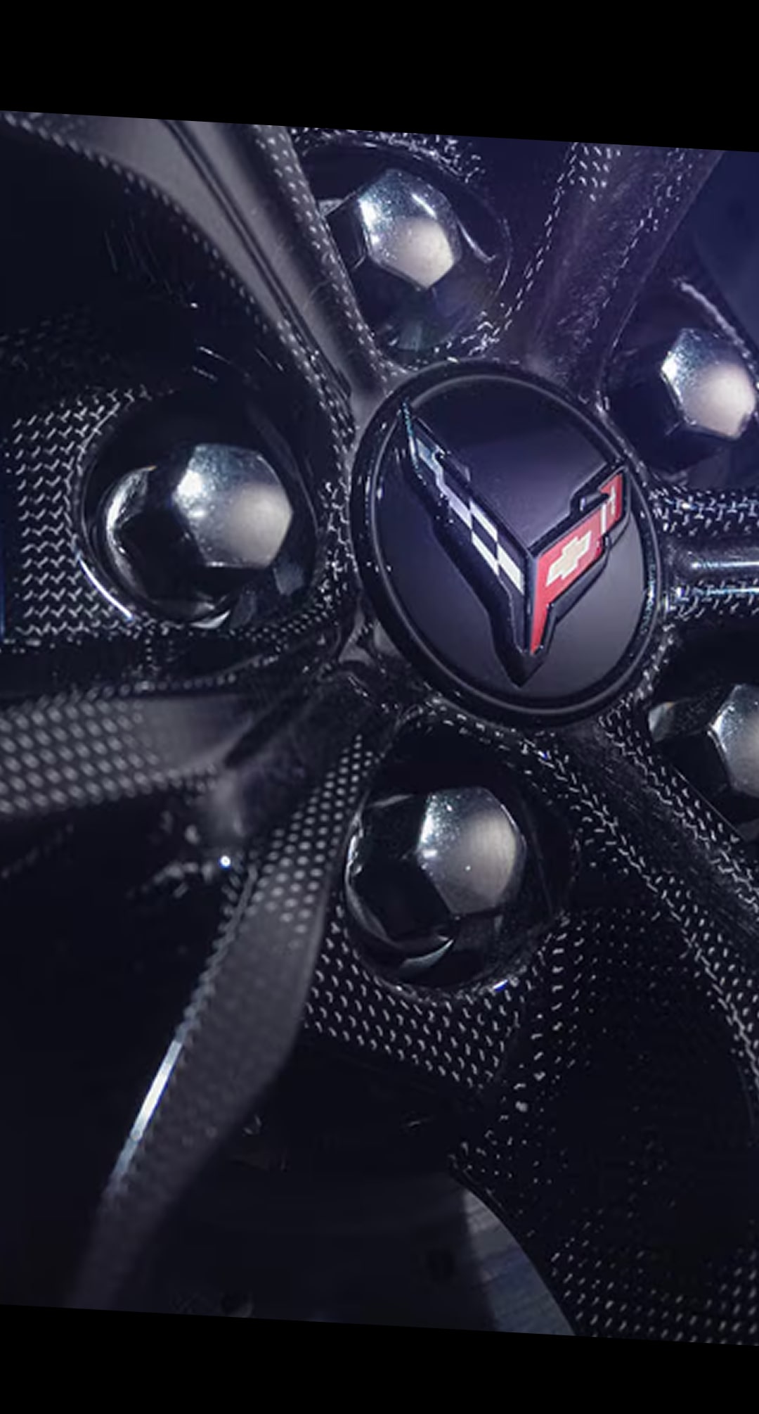 A Close-Up of a Black Carbon Fiber Performance Wheel Hub on the Chevy Corvette ZR1X Featuring a Corvette Crossed Flags Emblem at the Center Surrounded by Glossy Lug Nuts and Textured Spokes.
