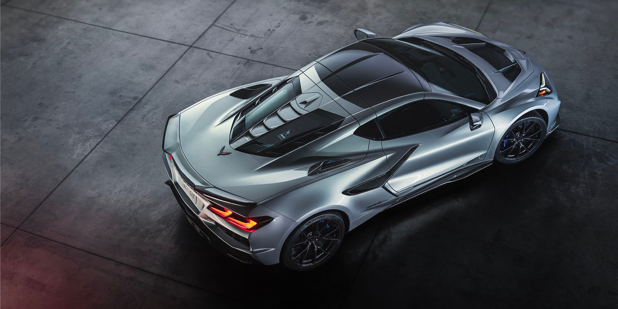 A Top-Down Three-Quarter View of a Silver Chevy Corvette ZR1X Showcasing Sculpted Aerodynamic Body Lines, a Vented Engine Cover, and Black Performance Wheels on a Dark Studio Floor.
