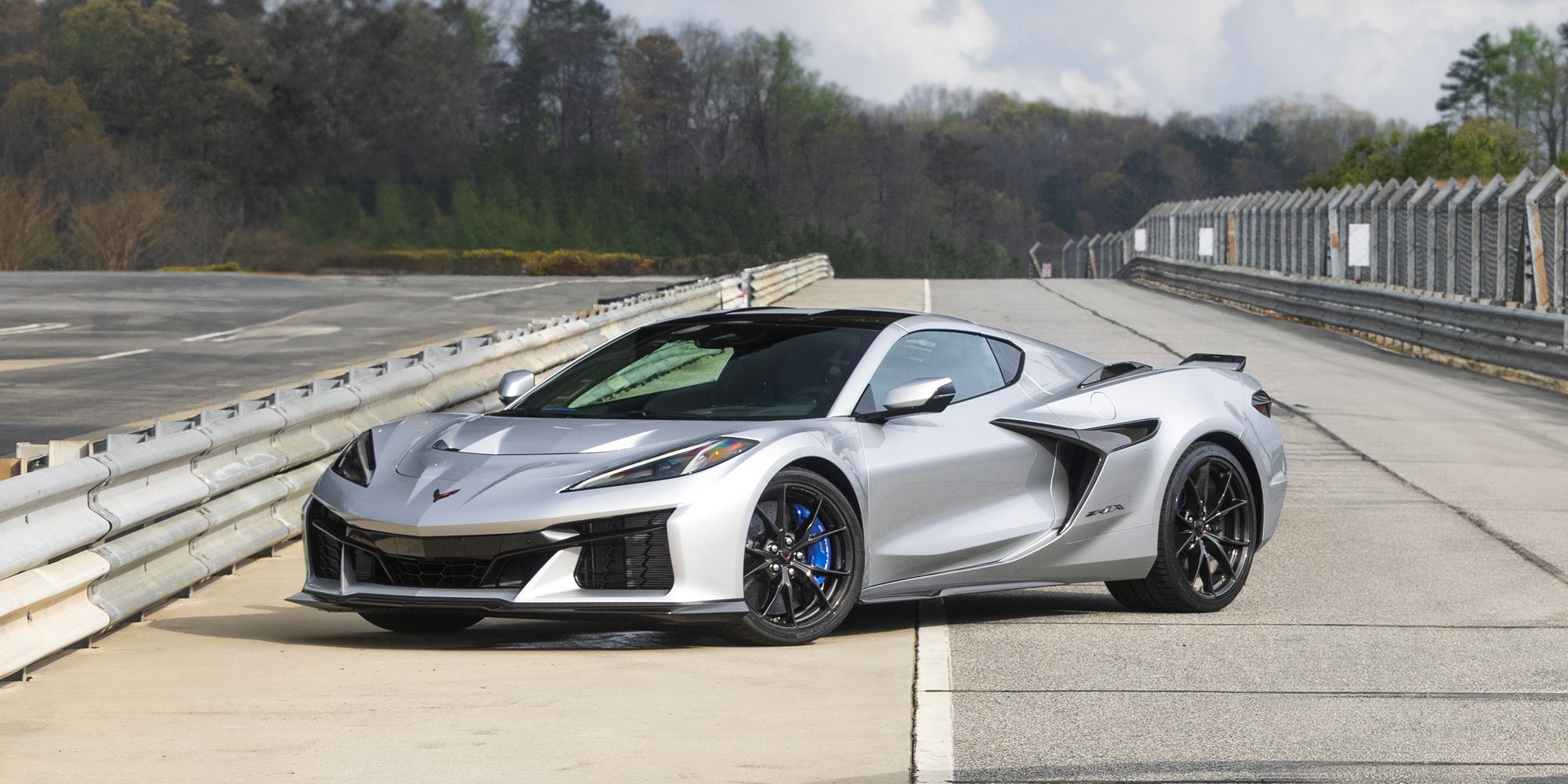A Silver Chevy Corvette ZR1X Driving on an Open Test Track With Black Wheels Surrounded by Guardrails and a Tree-Lined Background.