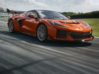 An Orange Chevy Corvette ZR1X Driving on a Paved Track with Smooth Body Lines, Low Profile Stance, and Green Grass Along the Roadside.