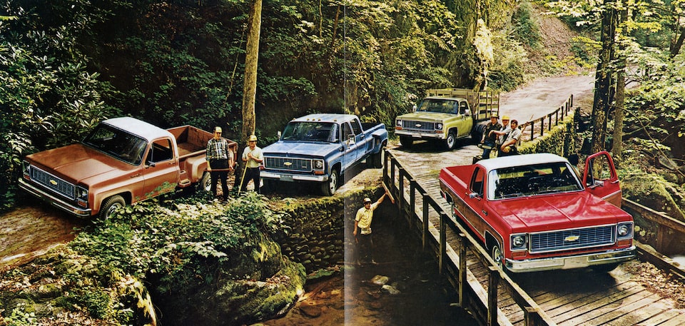 A photo from the original 1973 Chevy truck brochure showing four C/K Series pickups clustered together on a dirt road and a wooden bridge next to a steep, tree-covered hillside.
