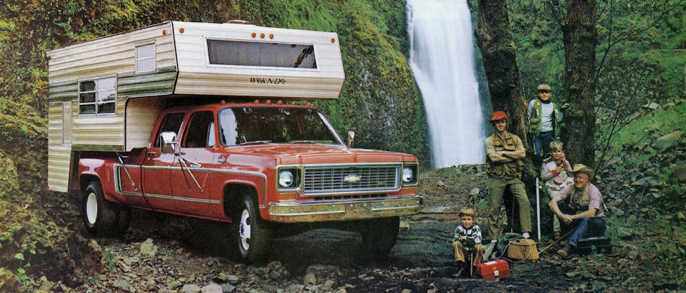 A photo from the original 1973 Chevy truck brochure showing a red C/K Series pickup with a bed camper, sitting in a wooded area near a waterfall. A man and four boys of different ages sit and stand next to the truck.