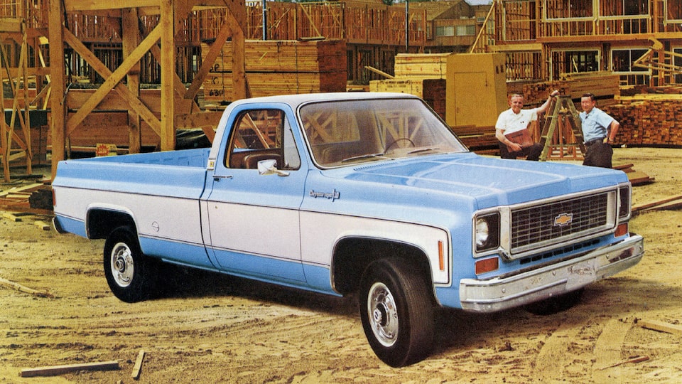 A photo from the original 1973 Chevy truck brochure showing a blue and white C/K Series pickup in a lumber yard. Two men stand behind the truck.