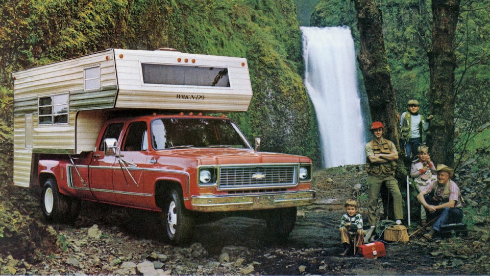 A photo from the original 1973 Chevy truck brochure showing a red C/K Series pickup with a bed camper, sitting in a wooded area near a waterfall. A man and four boys of different ages sit and stand next to the truck.