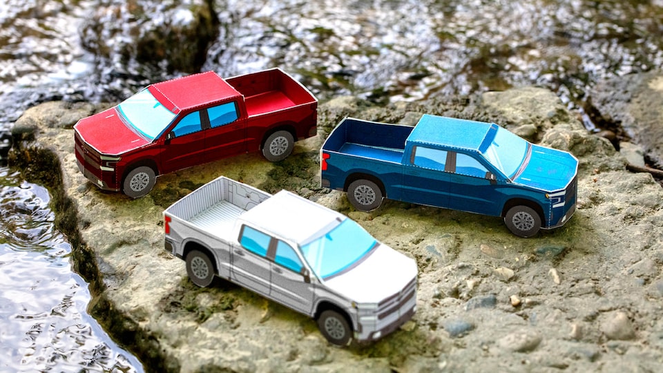 Miniature paper models of the 2020 Silverado RST Diesel in Cajun Red, Silver Ice Metallic, and Northsky Blue Metallic sitting on bare, rocky ground.