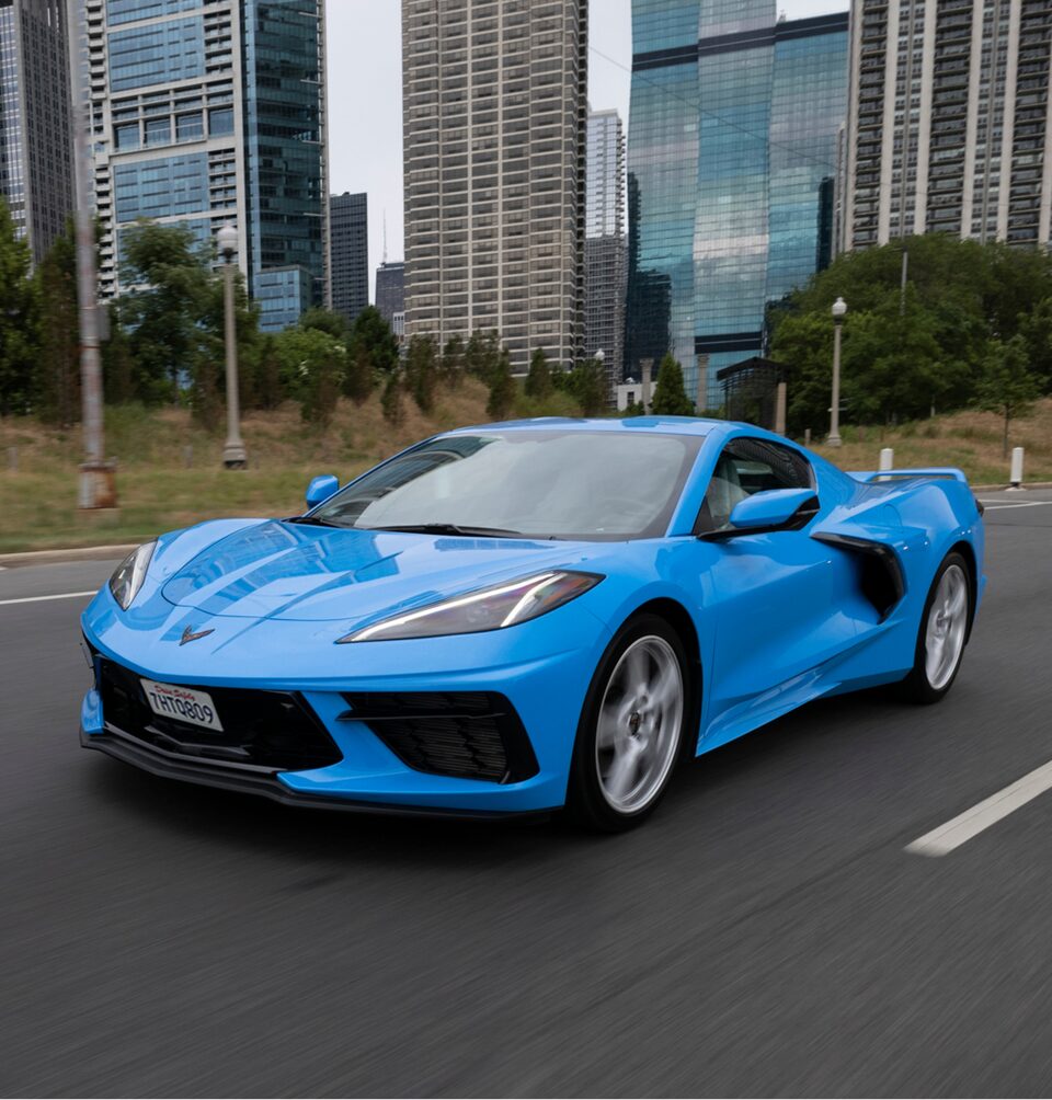 A bright blue Corvette drives on a road with skyscrapers in the background.