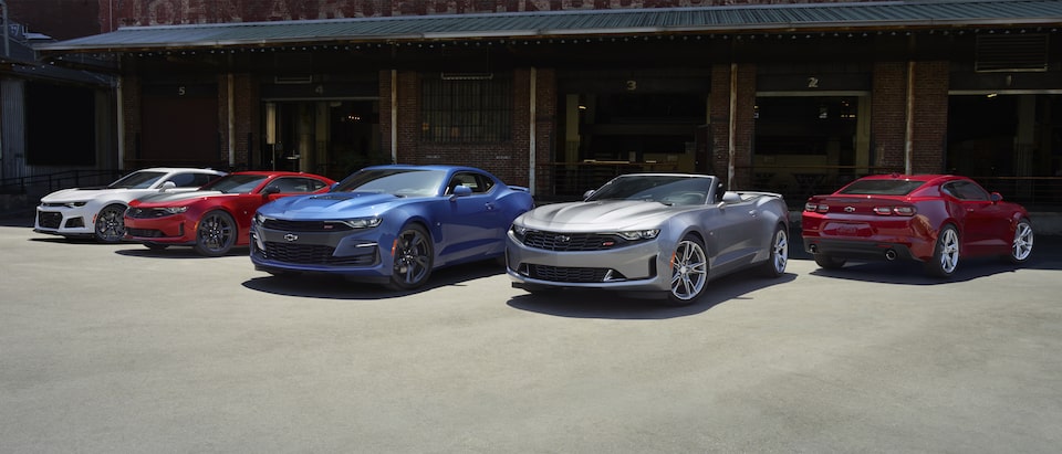 Un grupo de cinco Camaro estacionados en un muelle de carga: Coupes en color blanco, rojo y azul y un convertible en color plata.