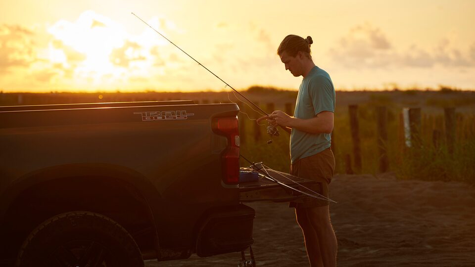 A man wearing a t-shirt and a backward baseball cap stands at the open tailgate of a Chevy Silverado holding a fishing pole, with the rising sun in the background.