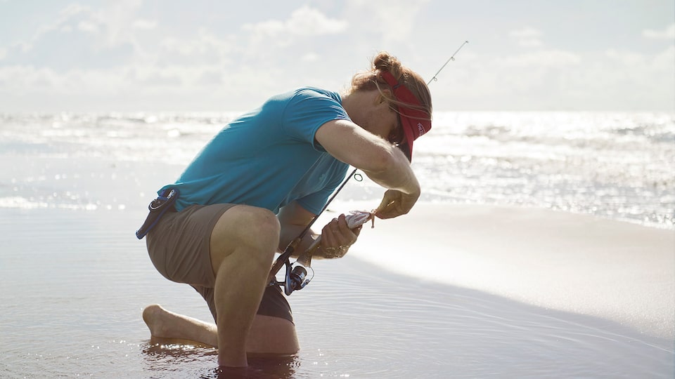 A man kneels on the sand at the ocean’s edge holding a fishing pole and baiting a hook.