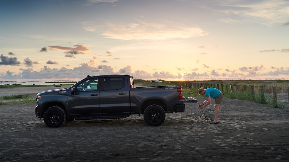 A man leans over to work on fishing poles that are resting against the open tailgate of a Chevy Silverado, with the rising sun in the background.