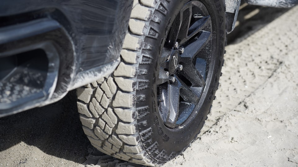 A close-up of the sand-covered tire of a Chevy Silverado as it drives along a beach area.