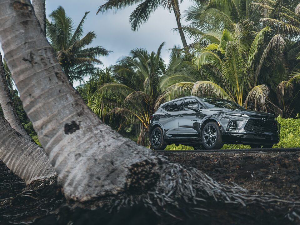 Una Chevy Blazer RS en Gris Hierro Metálico va por un camino en Hawái rodeado de vegetación y palmeras.