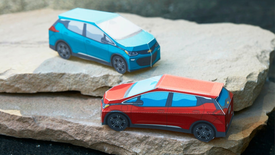 Miniature paper models of the Bolt EV in Cajun Red and Oasis Blue sit on a stack of flat rocks.