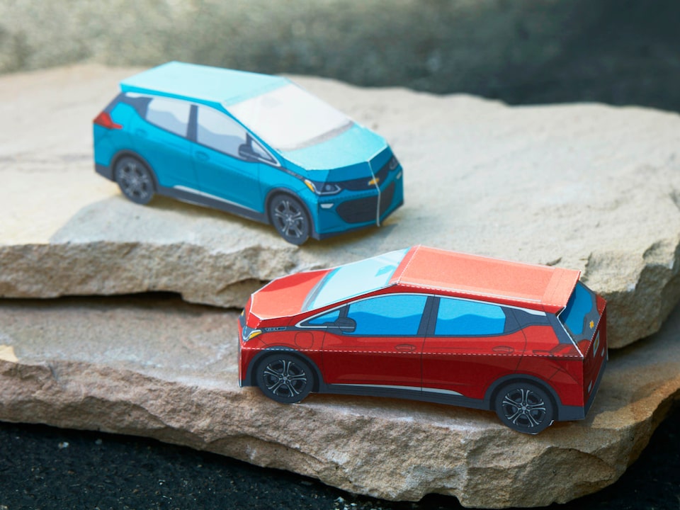 Miniature paper models of the Bolt EV in Cajun Red and Oasis Blue sit on a stack of flat rocks.