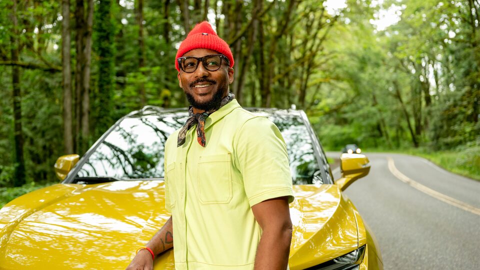 Redray Frazier, que tiene una camiseta amarilla y un sombrero rojo brillante, está parado delante de una Blazer de color amarillo brillante estacionada al costado de un camino rodeado de árboles.