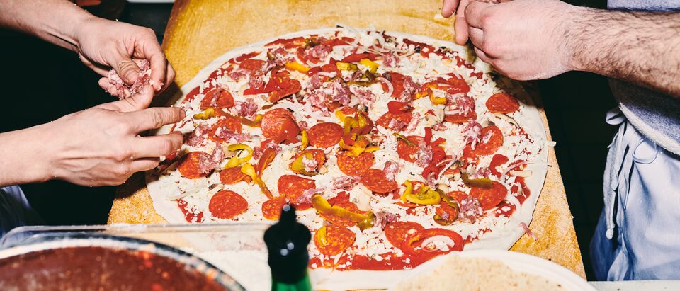 An overhead view of a pizza being made on a large wooden paddle, with two people’s hands adding toppings.