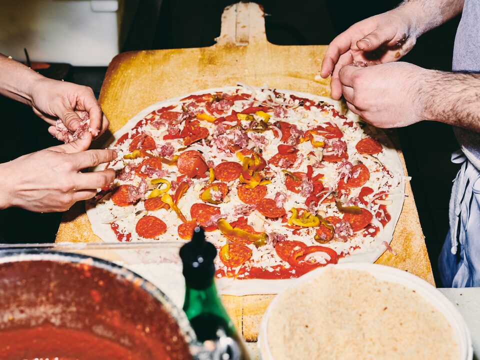 An overhead view of a pizza being made on a large wooden paddle, with two people’s hands adding toppings.