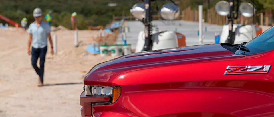 Vista lateral del frente de una Chevrolet Silverado roja. Jon Hagar camina hacia la camioneta y hay trabajadores de la construcción y equipos en el fondo.